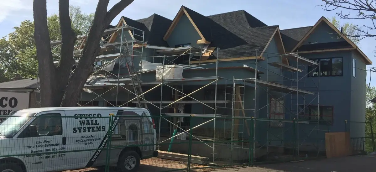 White Stucco Wall Systems van parked in front of a two-storey house wrapped in scaffolding, behind a green site fence