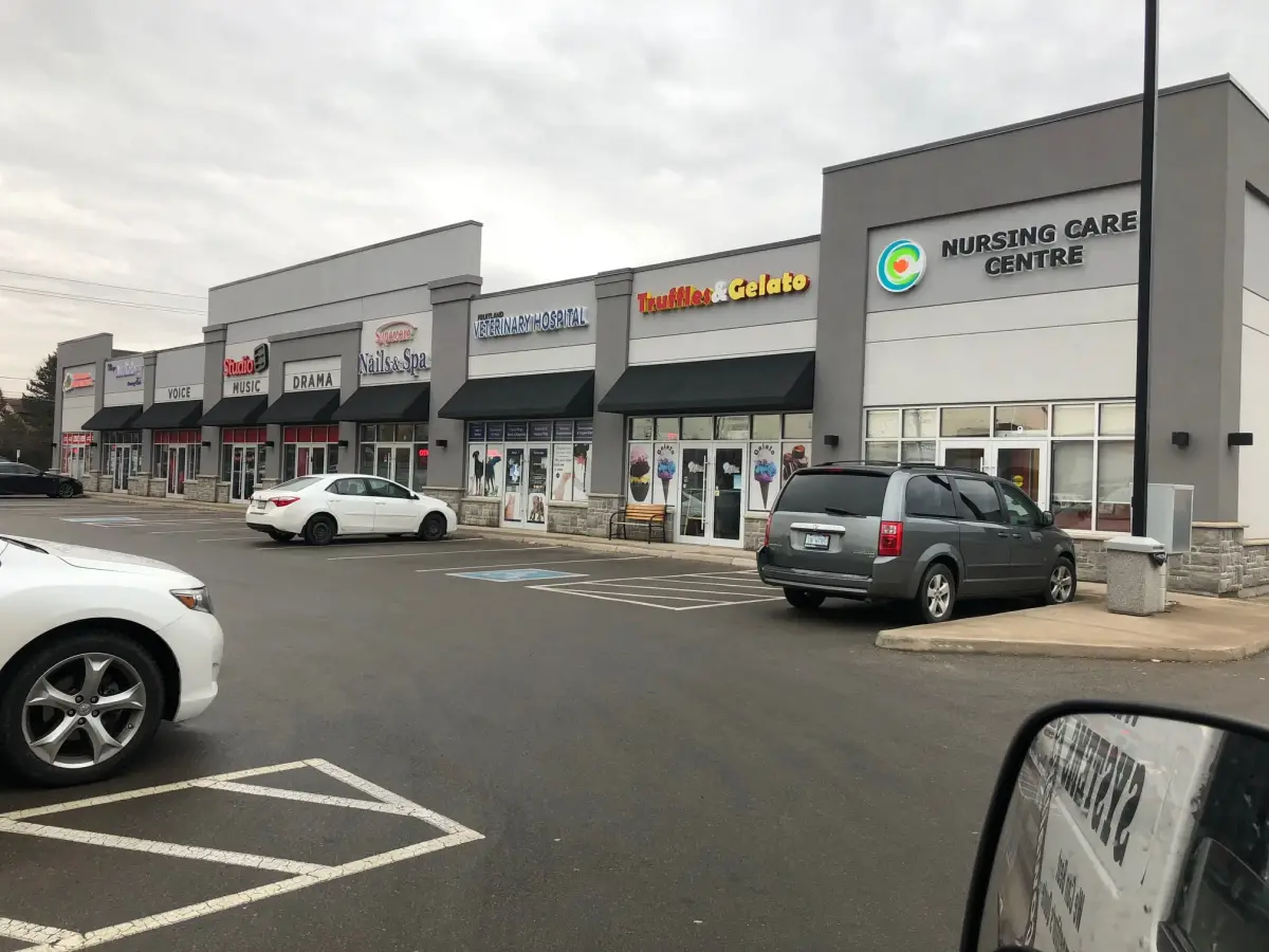 Single-storey retail plaza with grey facades, black awnings over the shop windows and a stone base along the walkway