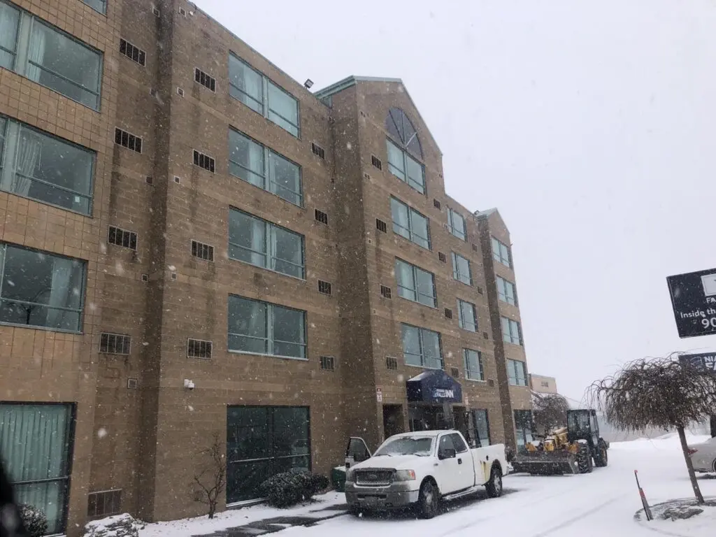 Five-storey tan building with rows of windows in falling snow, a white pickup truck parked at the entrance