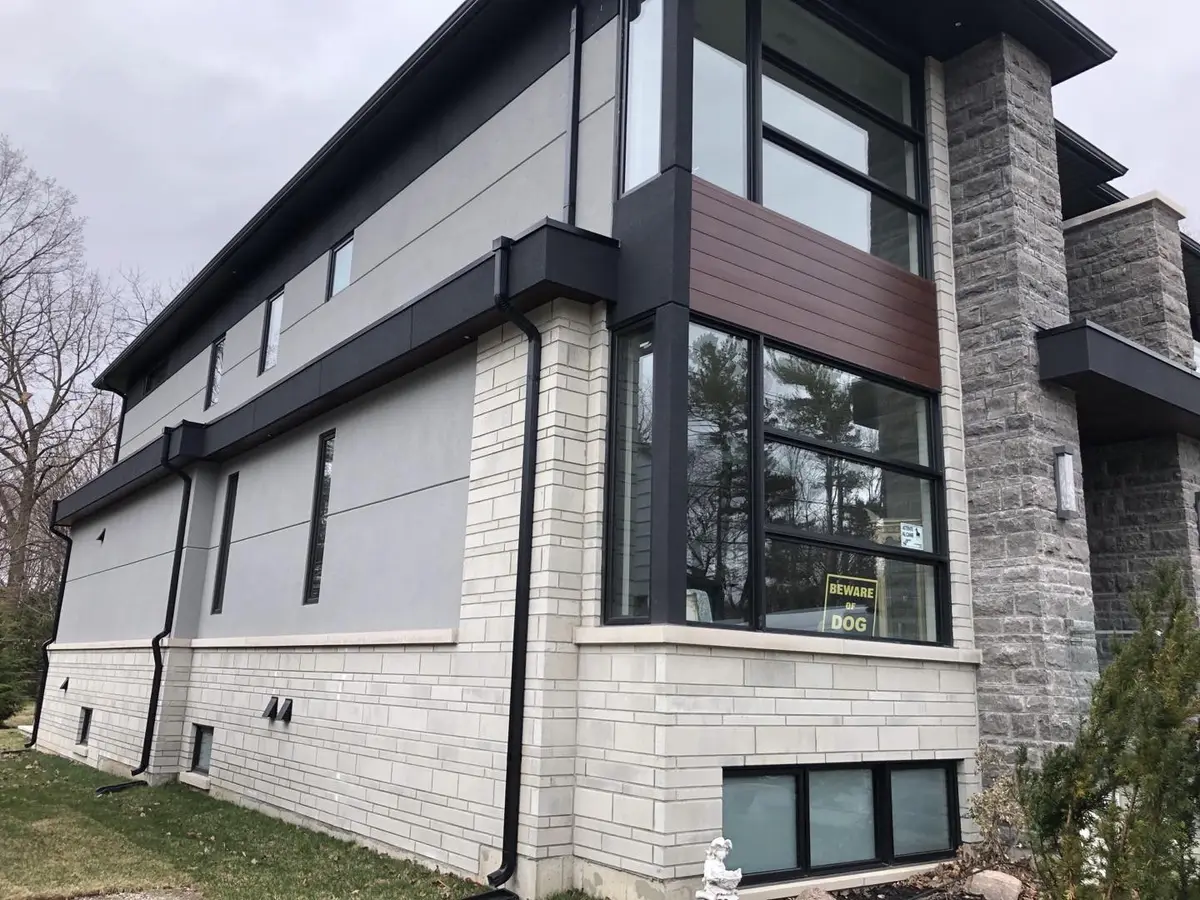 Modern two-storey house with grey stucco, a light stone base and stone columns, black eavestroughs along two roof edges and black downspouts