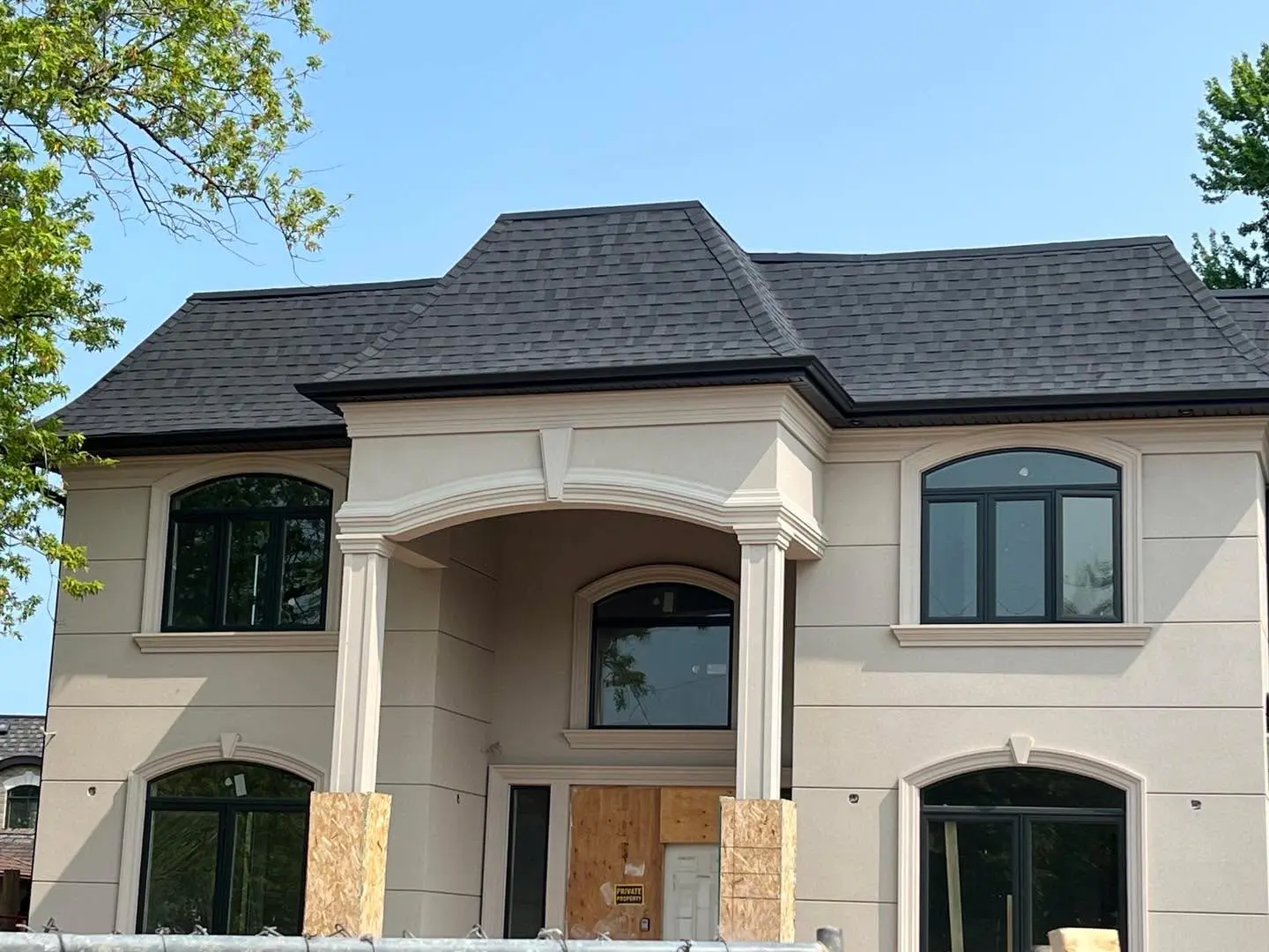 Cream stucco house with arched windows and a columned entrance under a dark shingle roof with a dark roof-edge line