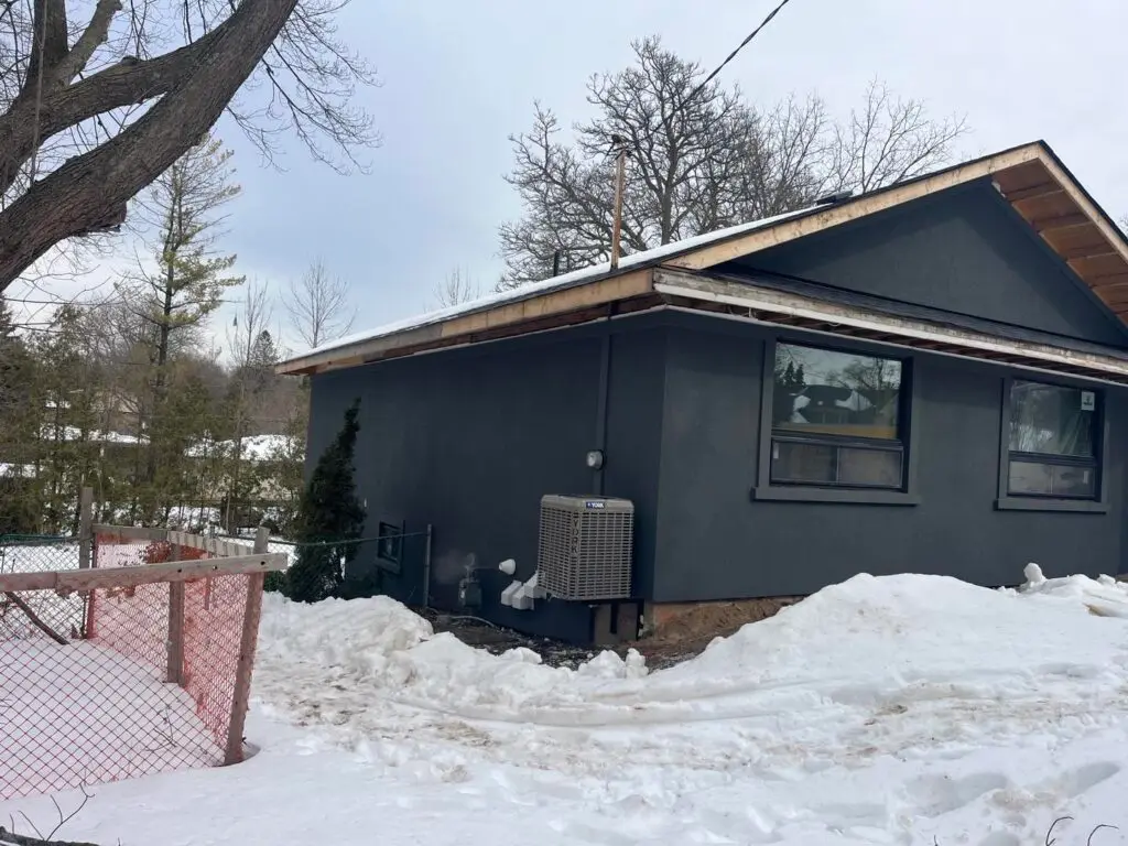 Charcoal stucco bungalow with bare wood fascia boards along the roof edge and deep snow in the yard
