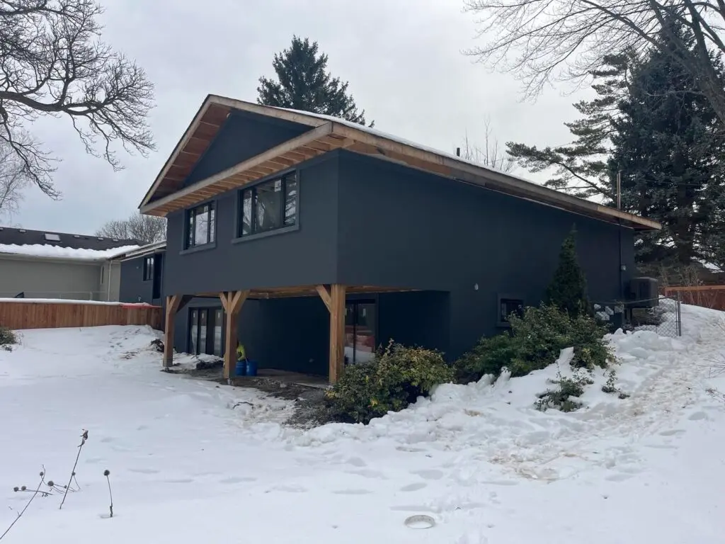 Raised bungalow finished in a dark charcoal stucco colour, with wood posts and snow on the lawn