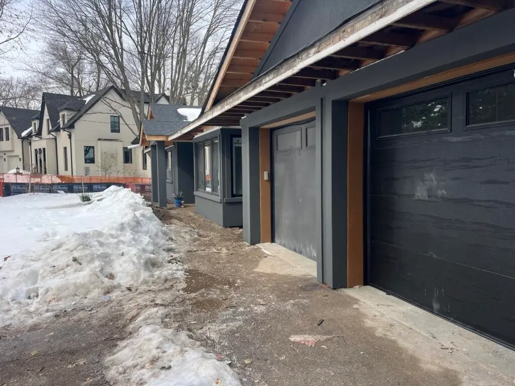 Open eave with exposed rafter ends above two charcoal garage doors, snow beside the driveway