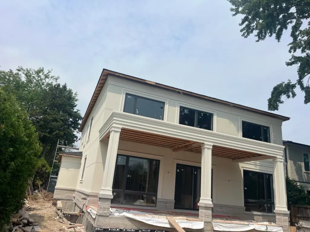 House mid-job with cream stucco walls and columns, stickers still on the windows, dimple membrane on the foundation and the soffit not yet closed